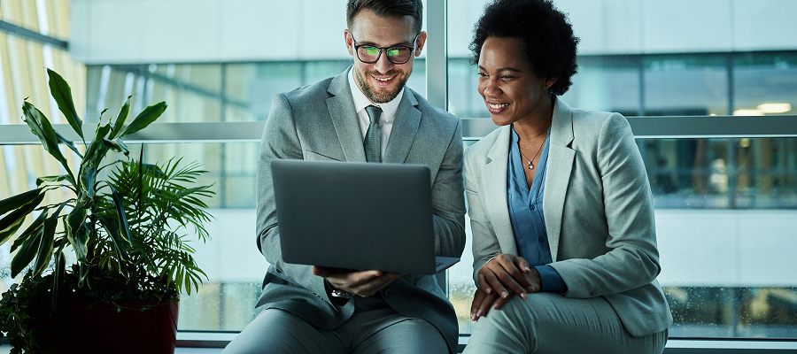 Man and woman in grey suits sitting on a ledge in an office building looking at a laptop