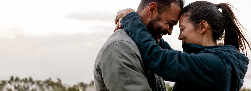 Man in grey jacket embracing and looking at woman in dark blue jacket under a grey sky