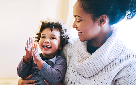 Woman in cream jumper sitting in front of a window and smiling at a toddler on her lap in a blue shirt